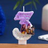 A purple glitter candle shaped like the number three, featuring small cartoon figures on the base.