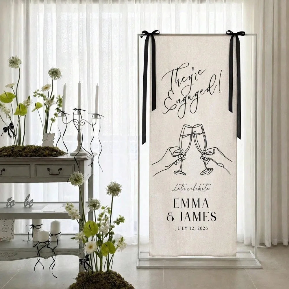 A vertical linen banner with black script text celebrating an engagement for Emma & James, displayed in a clear frame.