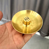 A round, brass incense stick holder plate with intricate patterns and nine small holes is held in a hand.