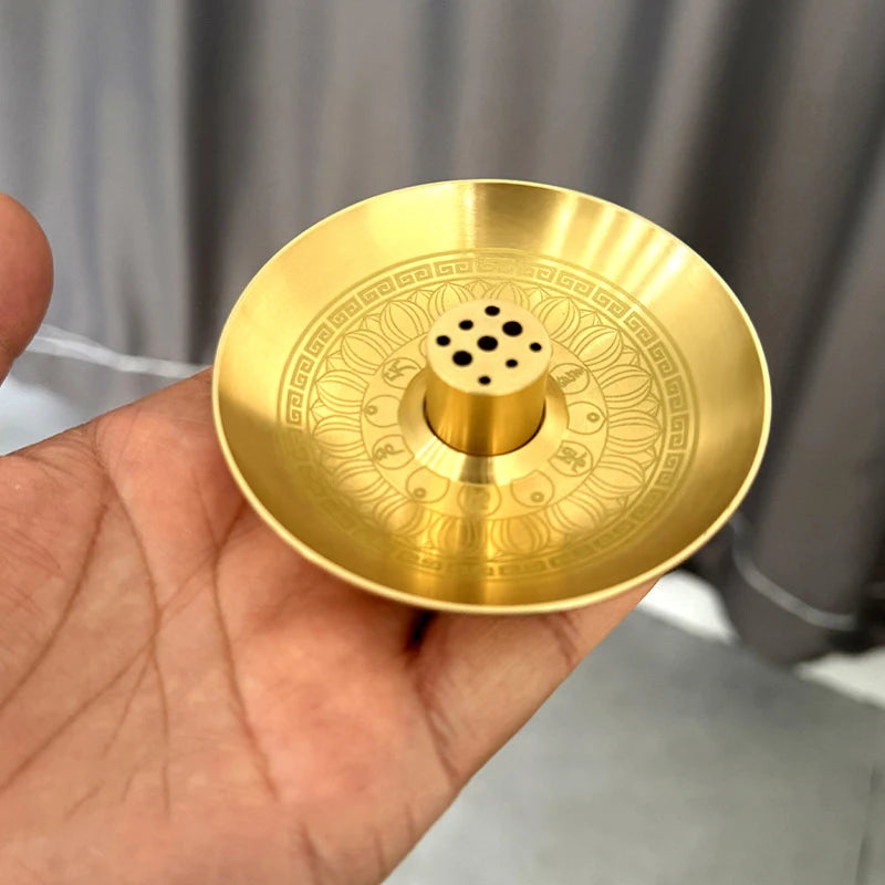 A round, brass incense stick holder plate with intricate patterns and nine small holes is held in a hand.