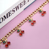 Gold chain necklace featuring red and green glass stone cherry charms on a pink background.