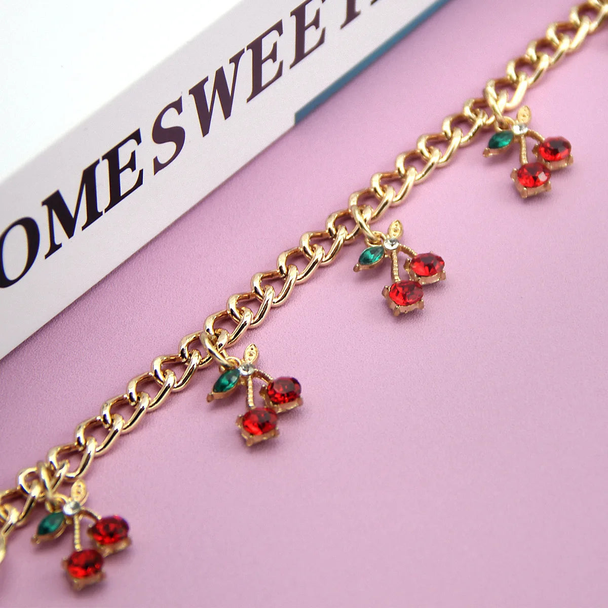 Gold chain necklace featuring red and green glass stone cherry charms on a pink background.