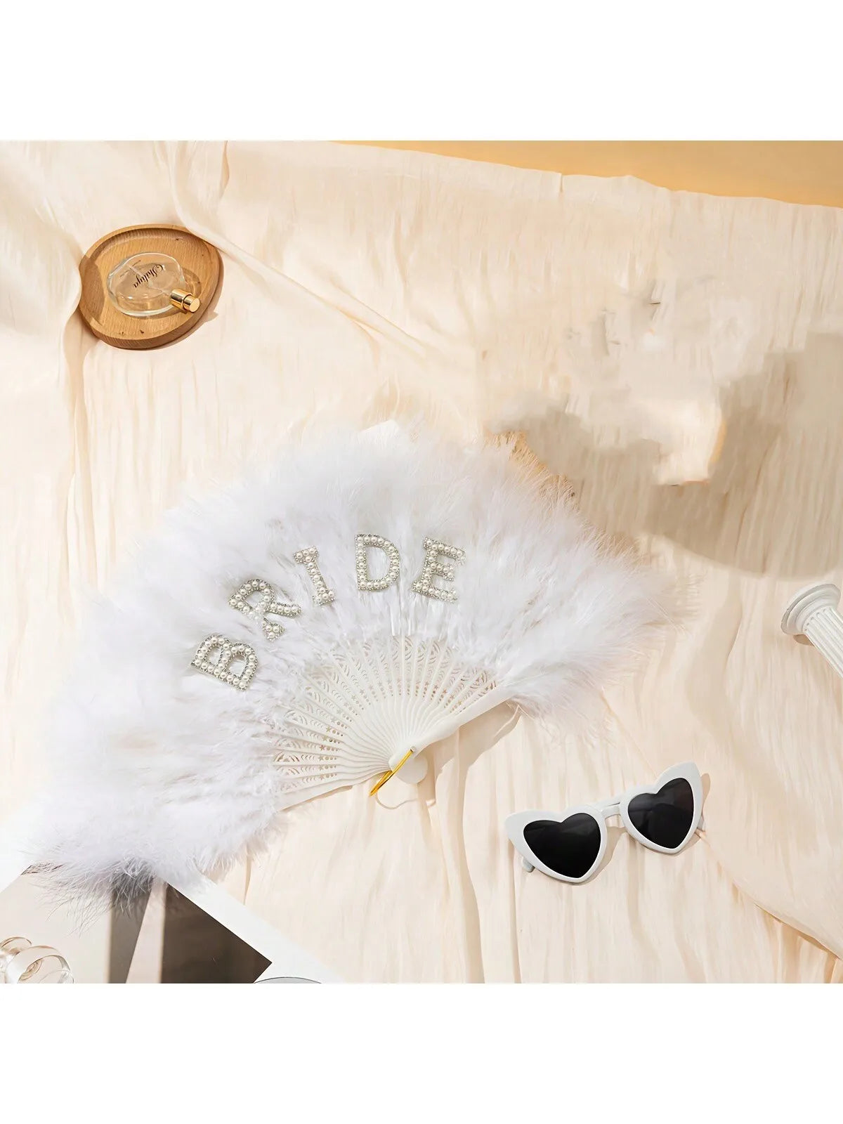 Bridal white feather hand fan with a rhinestone bride letter displayed on light fabric.