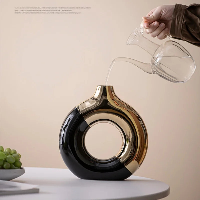 A hand pours water from a glass pitcher into a black and gold ceramic flower vase on a white table.