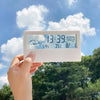 A hand holds a white LED digital thermometer and hygrometer displaying the time, date, weather icons, and temperature.