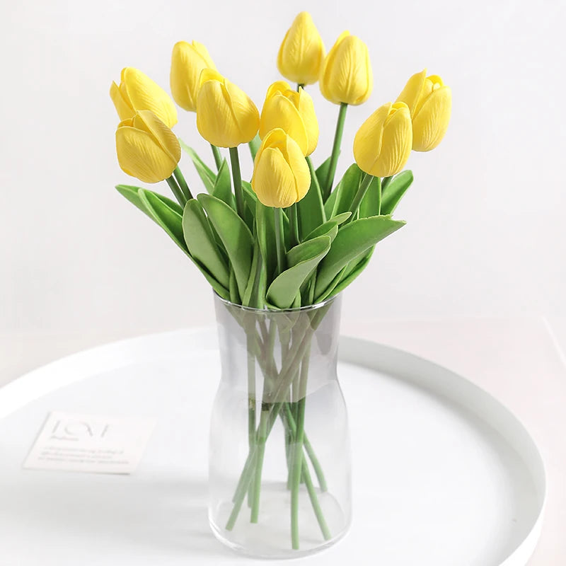 A bouquet of yellow artificial tulip flowers in a clear glass vase on a white surface.