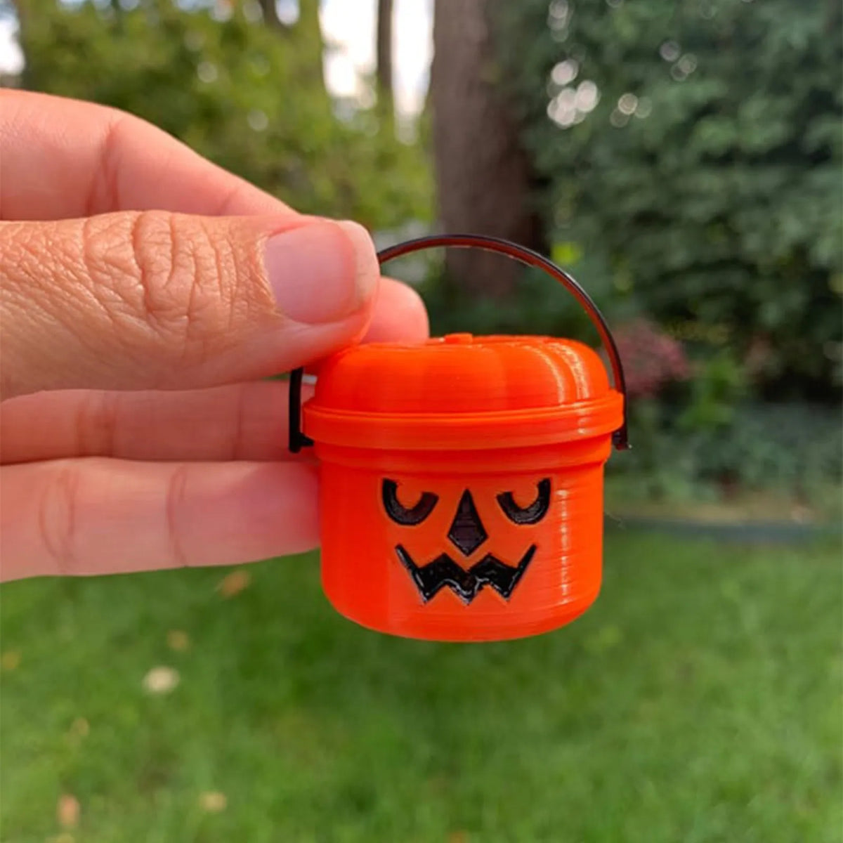 An orange miniature pumpkin Halloween bucket with a carved face is held in a hand outdoors.