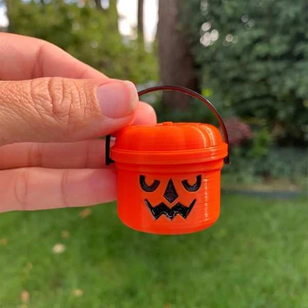 An orange miniature pumpkin Halloween bucket with a carved face is held in a hand outdoors.