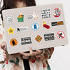 Una mano sostiene una laptop que muestra una colección de pegatinas y stickers de memes de dibujos animados sobre un fondo be