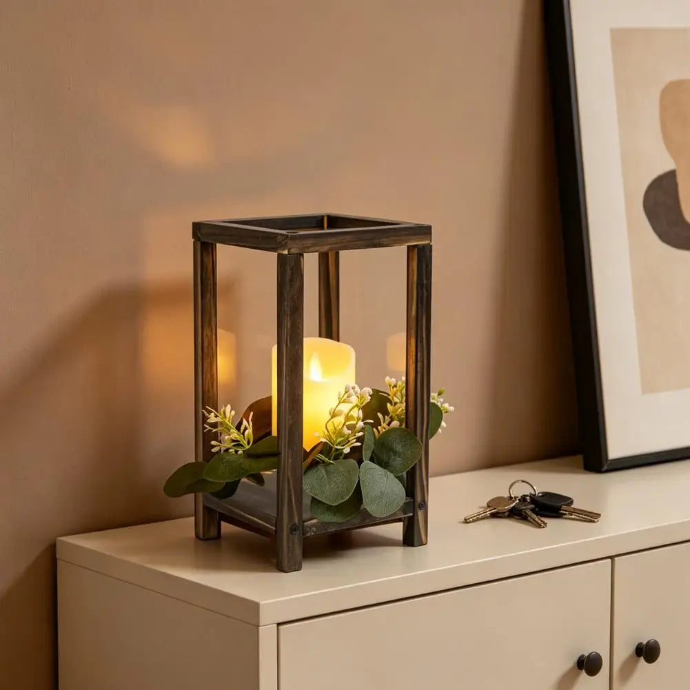A rustic wooden candle holder lantern with a yellow candle and green foliage sits on a light-colored cabinet.