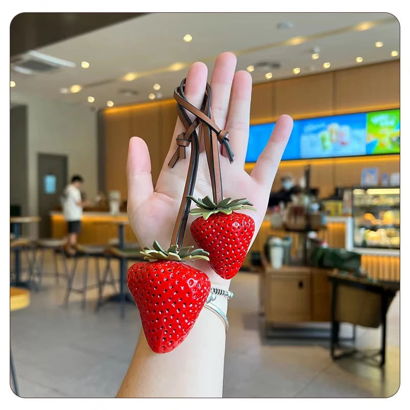 A hand holds a bright red strawberry charm and keychain with a leather strap against a blurred indoor background.