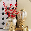 A golden ceramic vase with white stripes holds bright red berries and is displayed next to a gold geometric object.