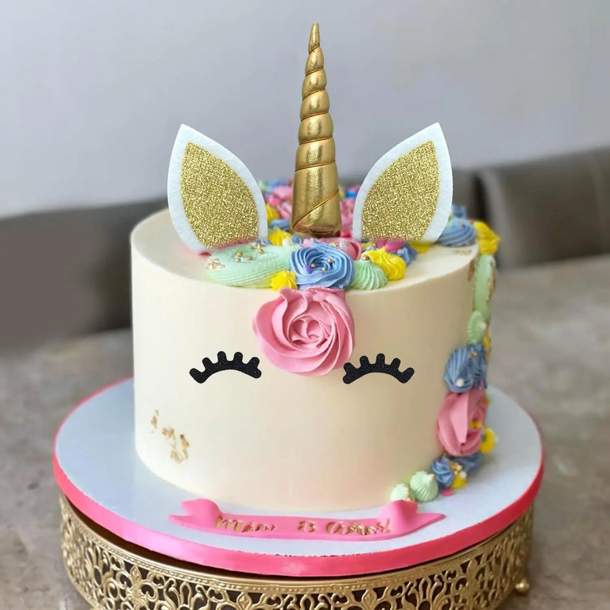 A light pink unicorn cake decorated with gold accents, white ears, and colorful floral elements.