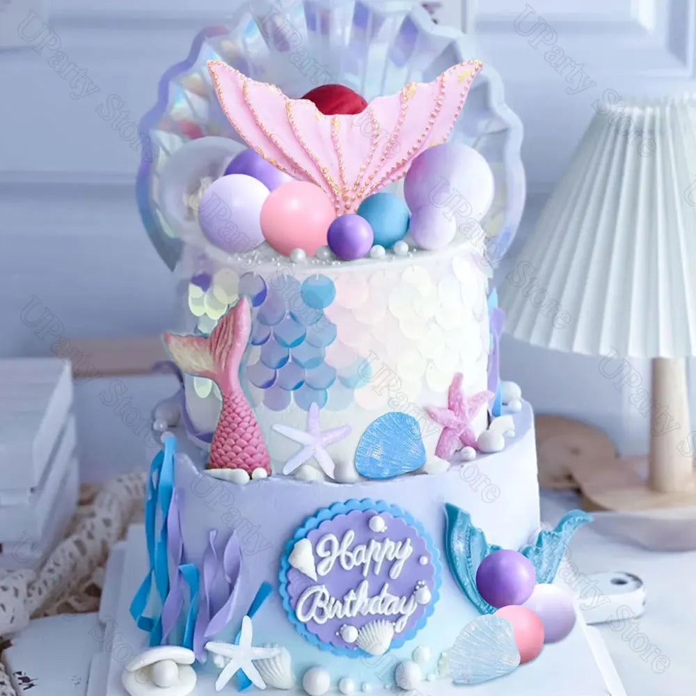 A tiered birthday cake decorated with mermaid themes, featuring pink and blue accents, seashells, and fish-like toppers.