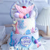 A tiered birthday cake decorated with mermaid themes, featuring pink and blue accents, seashells, and fish-like toppers.