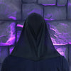 A person wearing a black hooded cloak stands in front of a purple, textured stone wall.