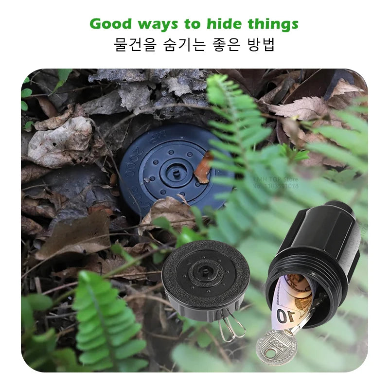 A black watering can key hider is partially hidden among leaves and dirt in an outdoor setting.