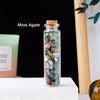 A glass bottle filled with various colored moss agate crystals displayed on a white surface.