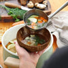 A hand holds a metal ladle over a dark brown coconut shell serving bowl filled with a clear broth and vegetables.