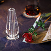 A handcrafted crystal glass rose vase with golden leaves sits next to a dark liquid in a glass.