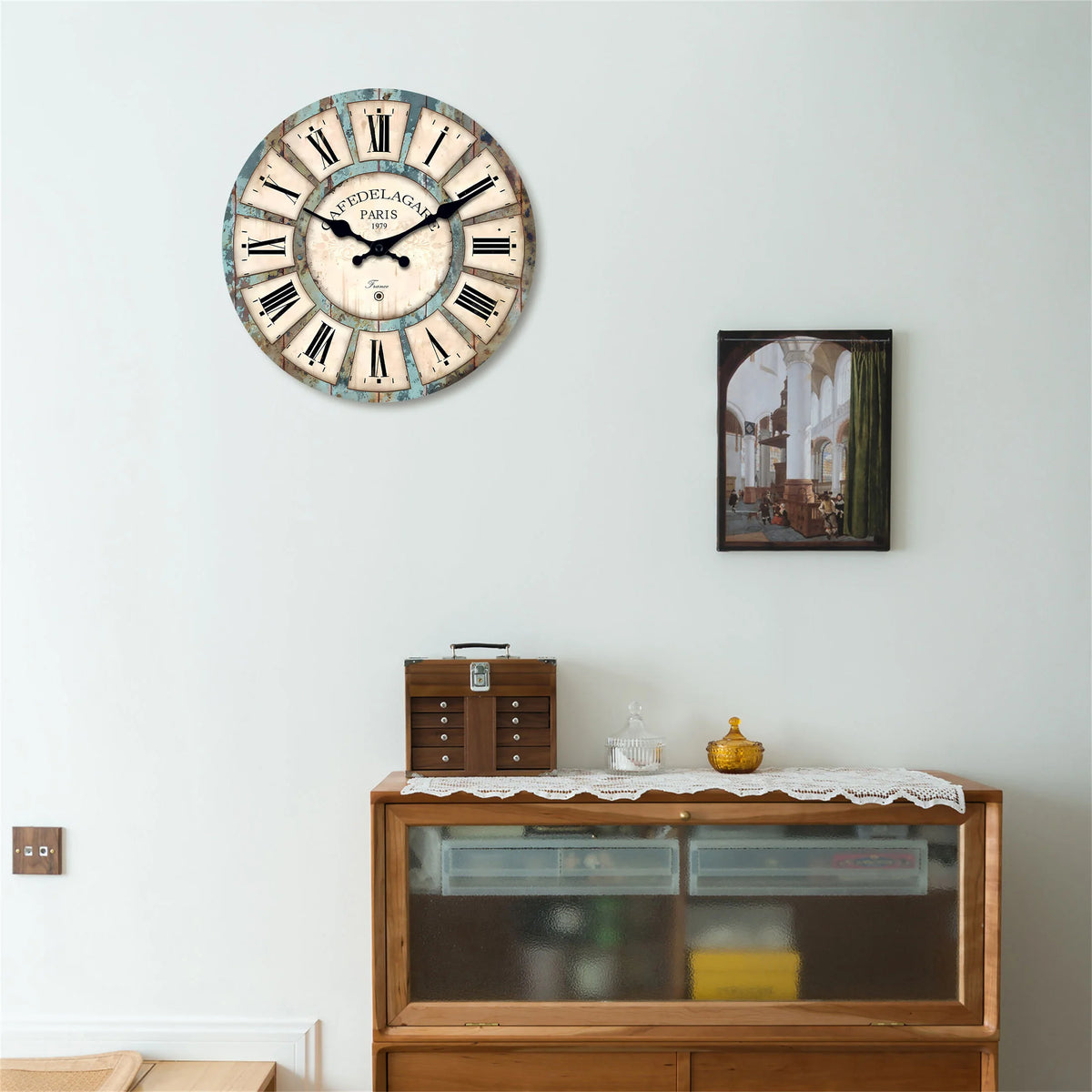 A rustic wooden wall clock with Roman numerals and a face showing Paris, mounted above a wooden cabinet.
