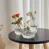 A set of three minimalist glass bud vases with delicate greenery and small white flowers displayed on a dark table.