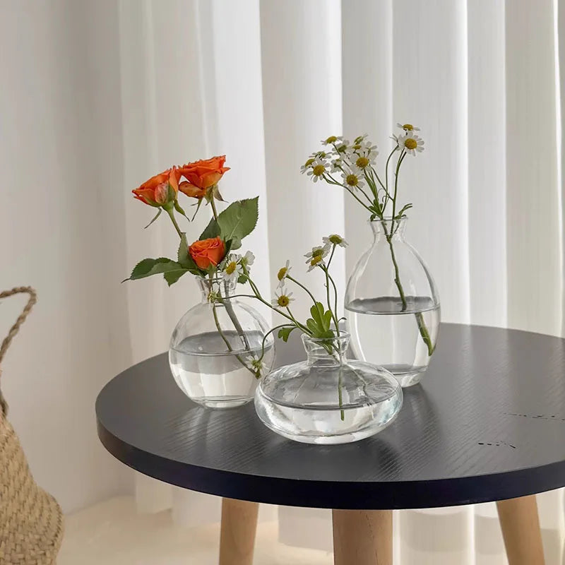 A set of three minimalist glass bud vases with delicate greenery and small white flowers displayed on a dark table.