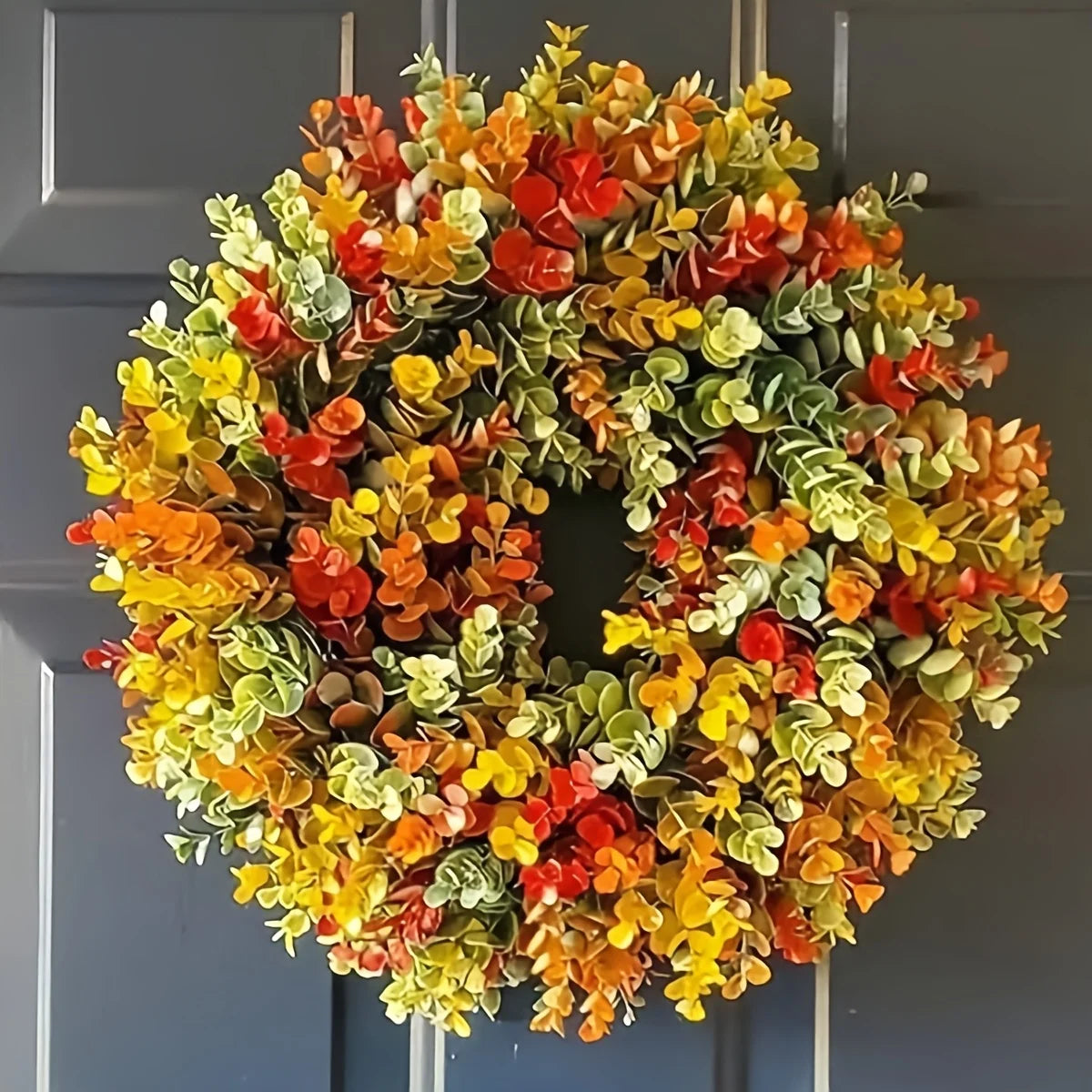 A circular wreath made of artificial flowers in shades of orange, yellow, and green hanging on a door.
