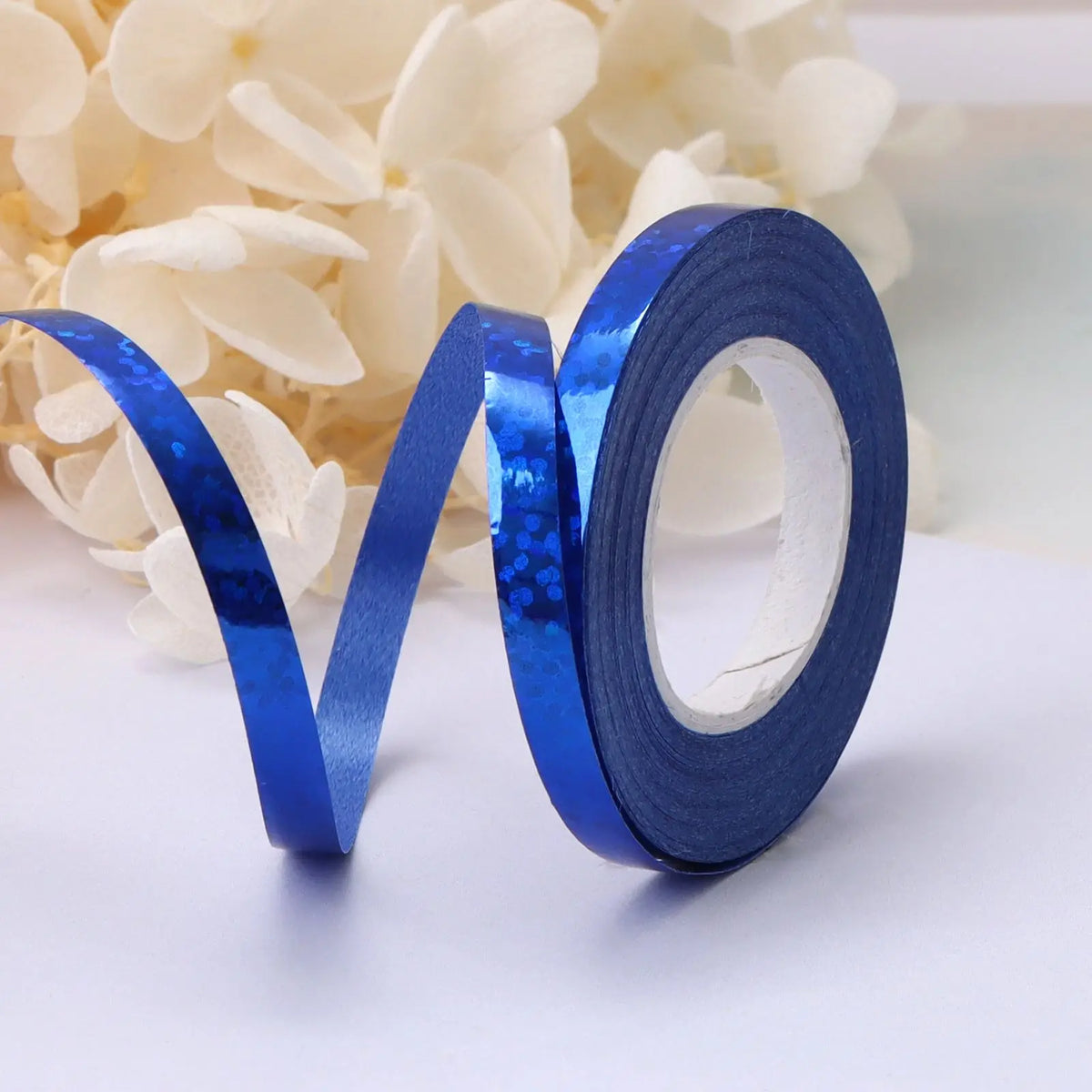 A roll of blue satin balloon ribbon lying on a white surface next to white flowers.