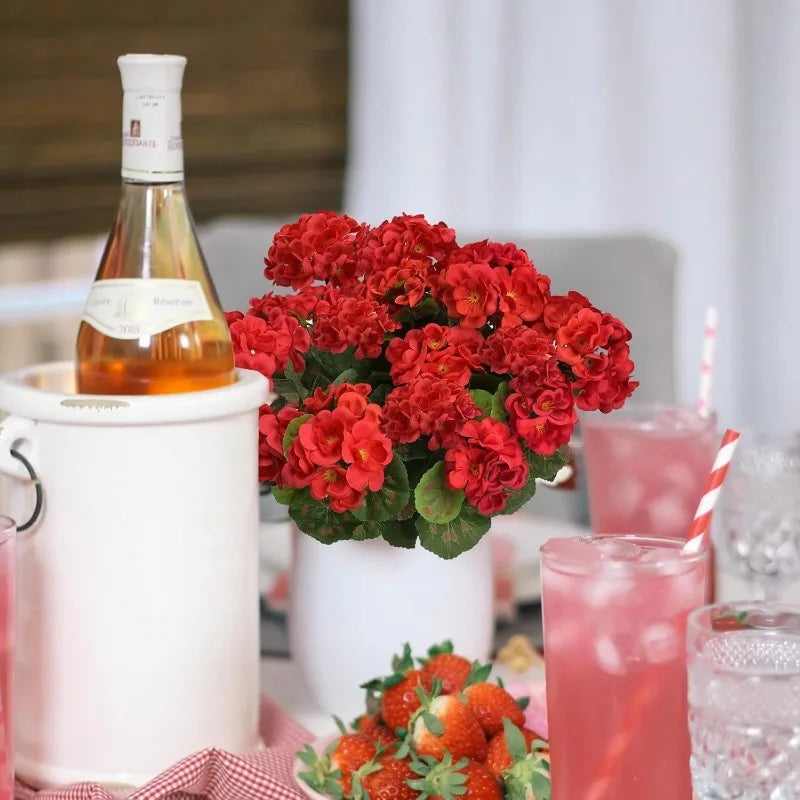 A bouquet of bright red artificial geranium flowers in a white pot next to a bottle of orange liquid and pink drinks.