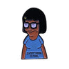 A enamel pin featuring a cartoon character with dark hair and glasses wearing a blue shirt.