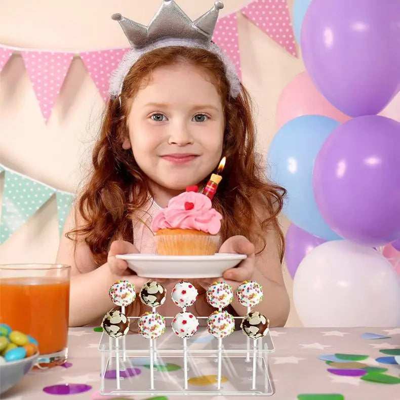 A young girl holds a pink cupcake and a display stand filled with colorful cake lollipops against a pastel background.