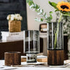 A tall glass vase with a wooden base holds a green leafy plant and an orange sunflower.
