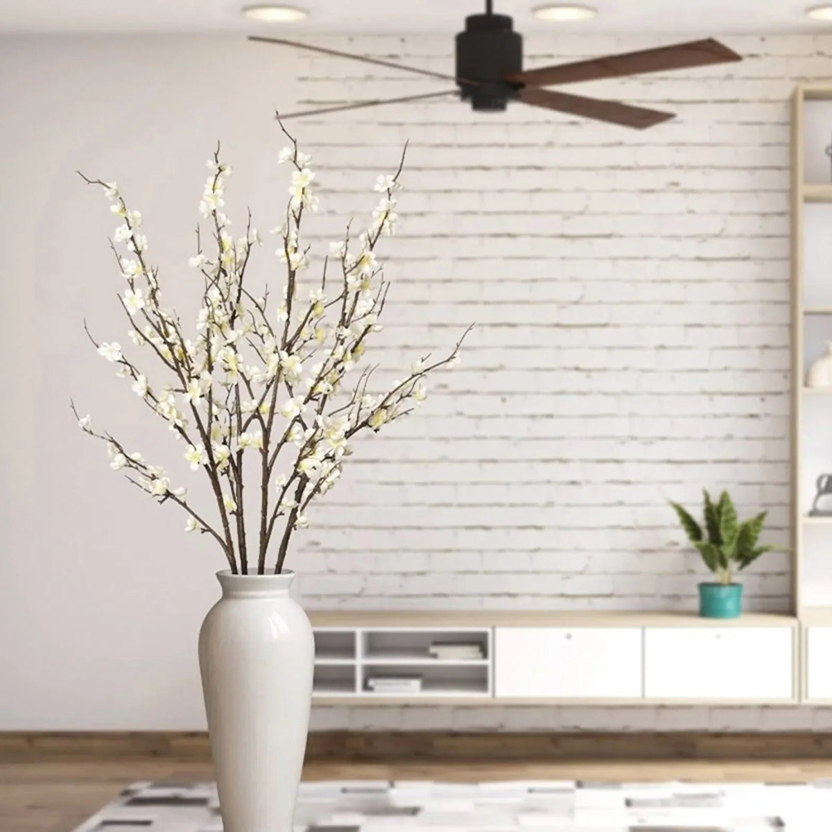 A vase holds a branch of white faux plum blossoms against a white brick wall.
