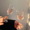 Two vintage French-style high-footed wine glasses filled with pink liquid are held against a light gray wall.