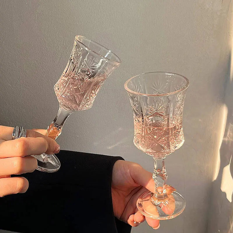 Two vintage French-style high-footed wine glasses filled with pink liquid are held against a light gray wall.