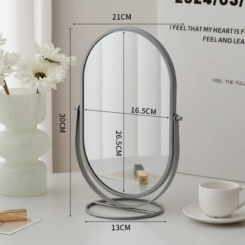 An elegant rotating oval vanity mirror with a silver frame and round base is displayed on a white surface.