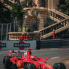 A red Formula 1 race car is speeding on a track, with grandstands and architecture in the background.
