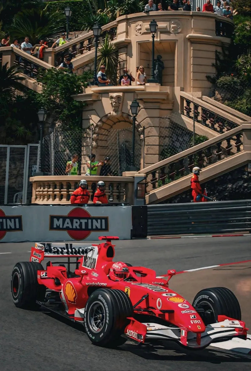 A red Formula 1 race car is speeding on a track, with grandstands and architecture in the background.