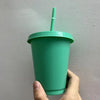 A hand holds a green plastic cold cup with a lid and straw against a light gray wall.