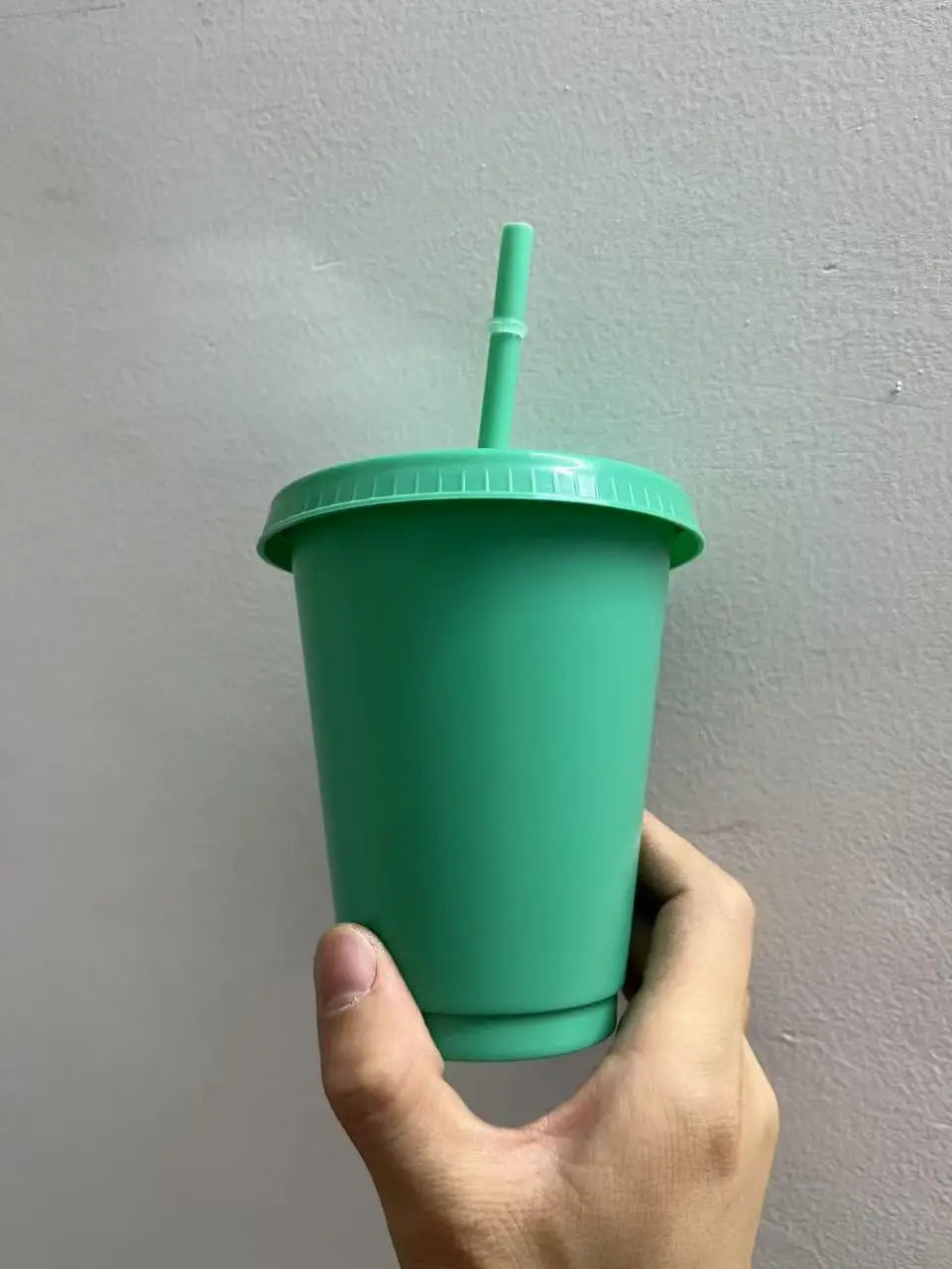 A hand holds a green plastic cold cup with a lid and straw against a light gray wall.