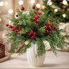 A bouquet of artificial Christmas berry stems and pine branches in a white vase on a wooden surface.