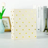 A square beige photo album with gold foil hearts on the cover, next to a white tulip and books.