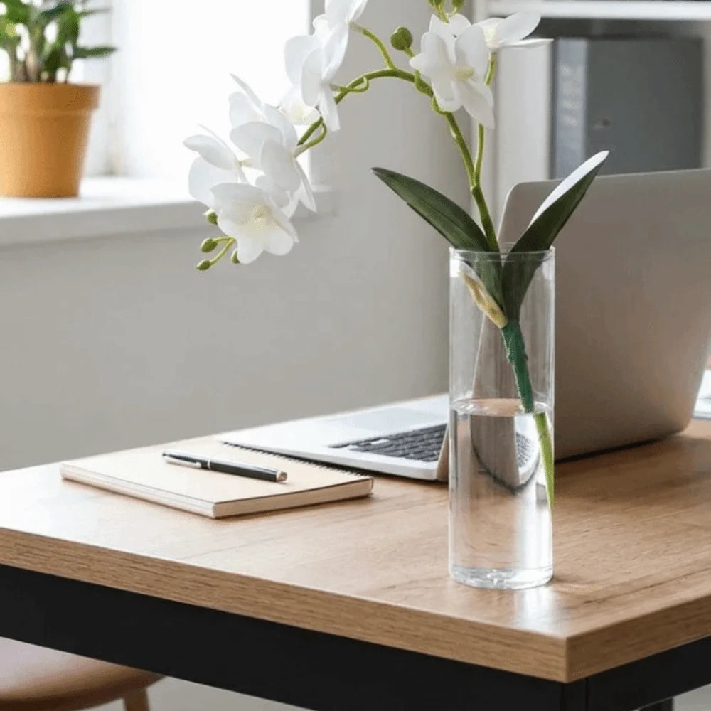 A clear glass cylinder vase holds white orchids on a wooden desk next to a laptop.