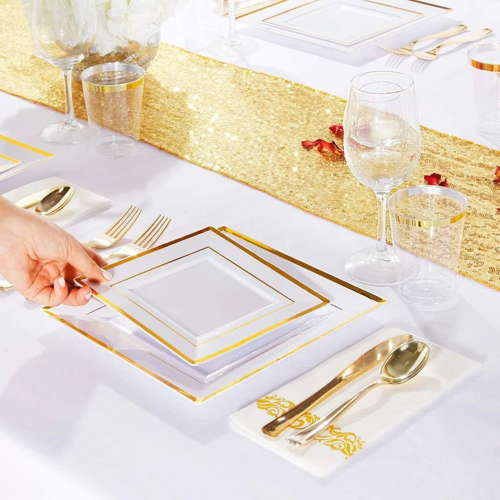 A close-up of a gold plastic dinnerware set laid out on a white tablecloth with gold glitter runner and glassware.
