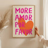 A framed canvas print with bright pink text reading MORE AMOR POR FAVOR on an orange heart, displayed on a light-colored wall