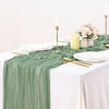 Sage green gauze table runner set with white plates, gold cutlery, and pink floral centerpiece.