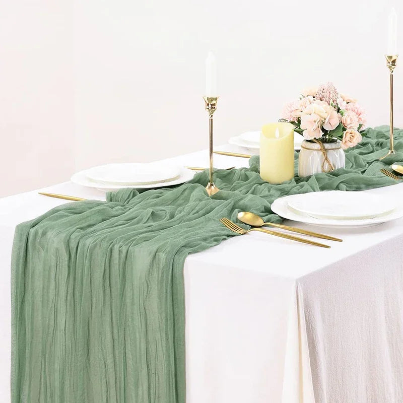 Sage green gauze table runner set with white plates, gold cutlery, and pink floral centerpiece.