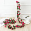 A long garland of pink roses with green leaves draped against a white wall and a white vase.