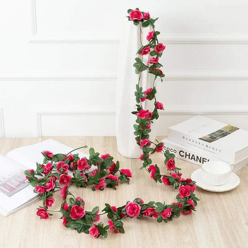 A long garland of pink roses with green leaves draped against a white wall and a white vase.